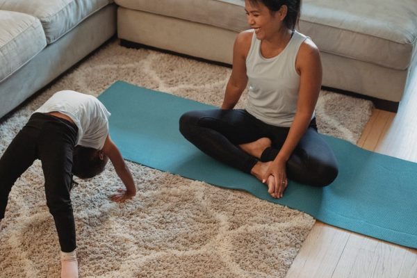 From above of happy mother sitting in lotus pose and looking at daughter bending over backwards at home in daytime