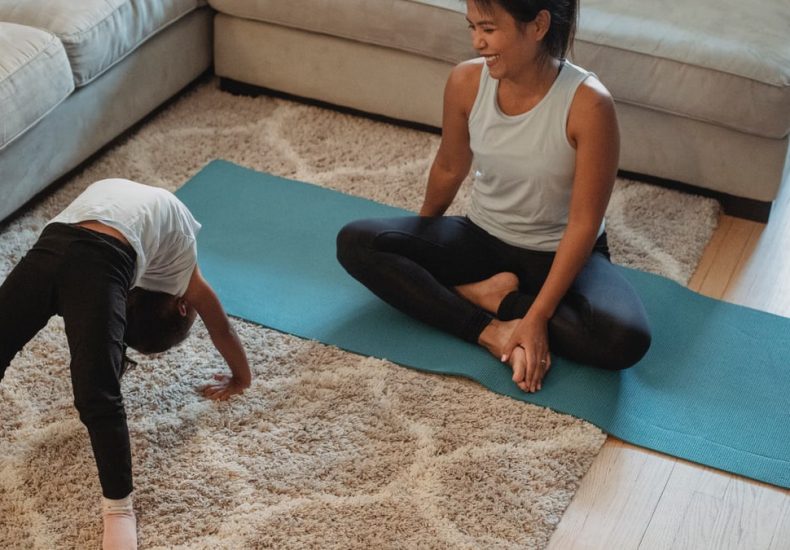 From above of happy mother sitting in lotus pose and looking at daughter bending over backwards at home in daytime