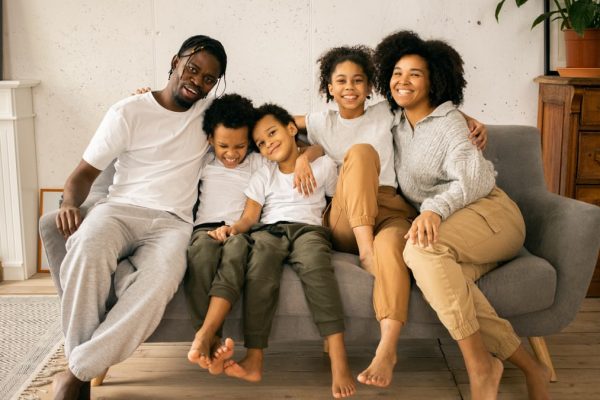 Full body of cheerful African American parents with children looking at camera while sitting on sofa in room with fireplace