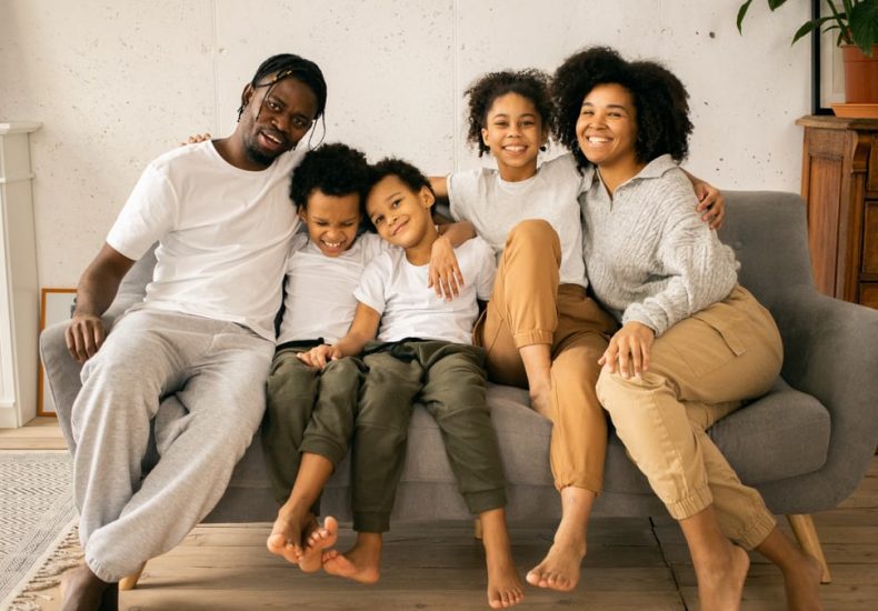 Full body of cheerful African American parents with children looking at camera while sitting on sofa in room with fireplace